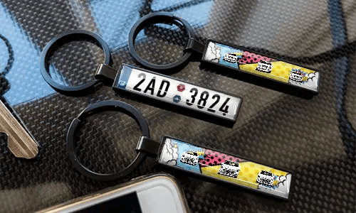 Sleutelhangers met kentekenplaat in stripstijl, weergegeven op een koolstoftextuur met kleurrijke autodesigns