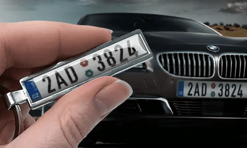 Portachiavi a forma di targa in stile fumetto con numero personalizzato, tenuto in mano vicino alla targa dell’auto corrispondente