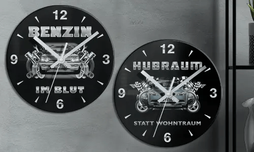 Zwei schwarze Wanduhren mit Autoillustrationen und deutschem Text: BENZIN IM BLUT und HUBRAUM STATT WOHNTRAUM